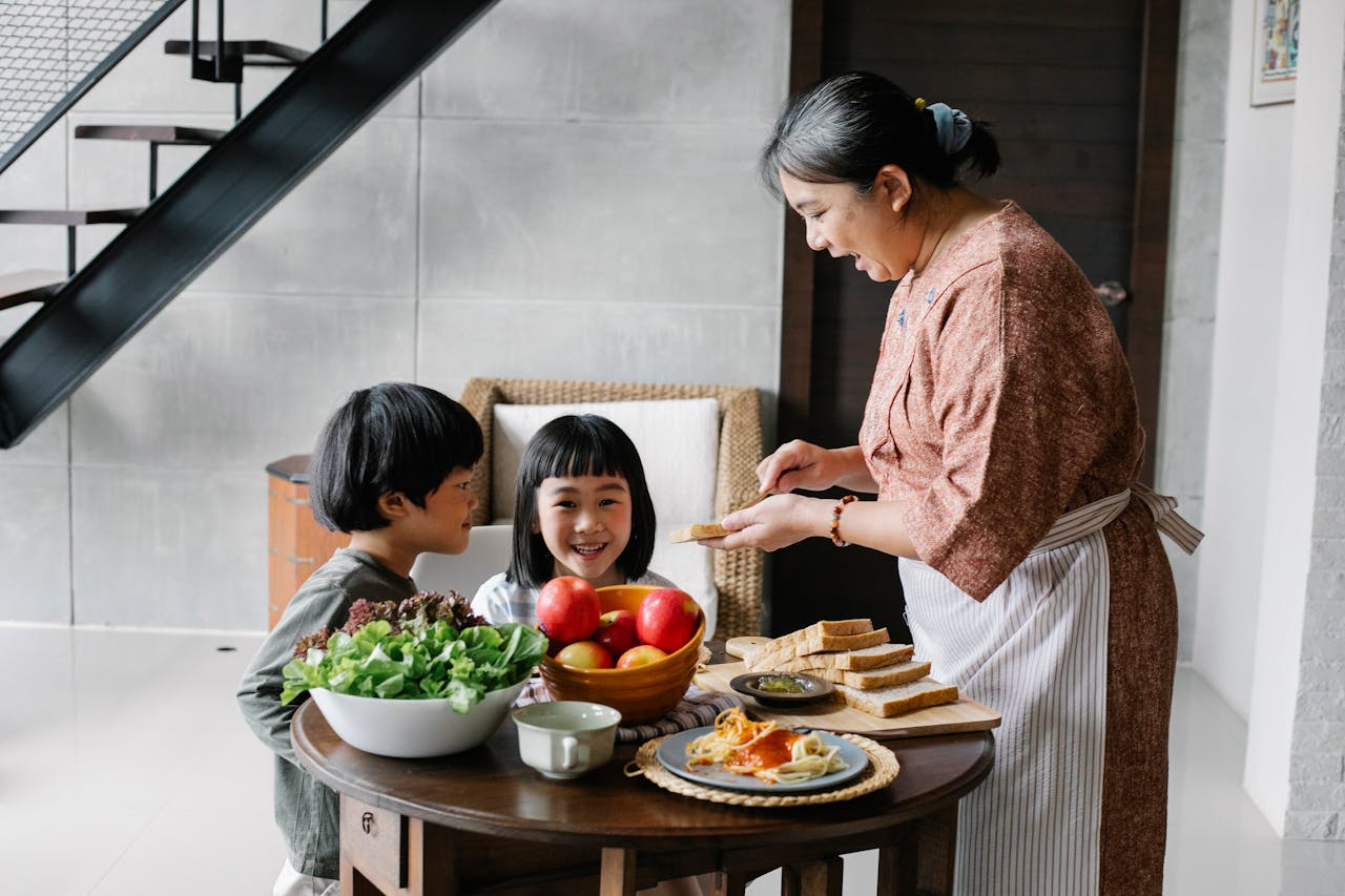 about-07 Side view of positive senior Asian female in apron spreading butter on bread while preparing breakfast for funny little grandchildren sitting at round table in cozy kitchen