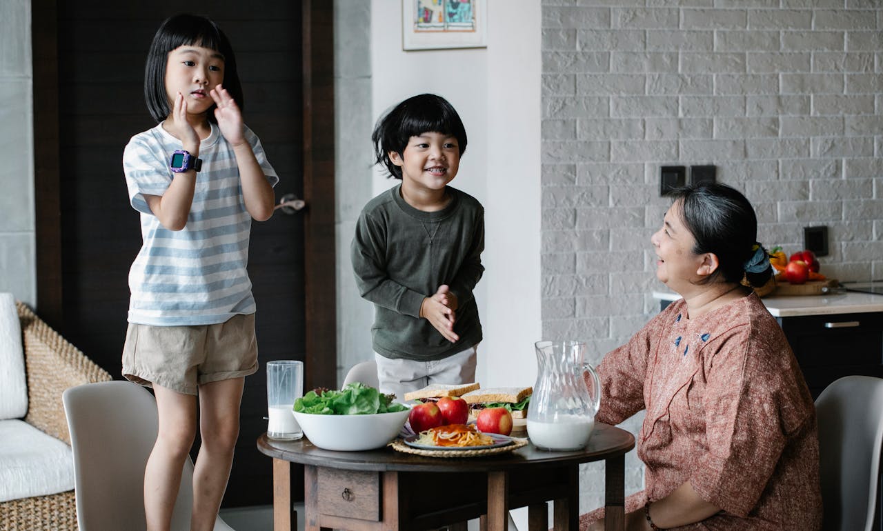 services-09 Asian grandmother sitting at served table during breakfast in kitchen while looking at Asian boy and girl having fun together