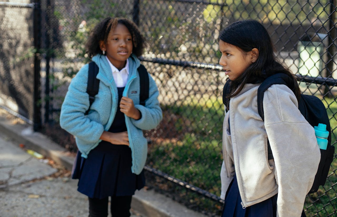 mission-02 Two diverse schoolgirls in warm clothing chatting outdoors on a sunny autumn day.