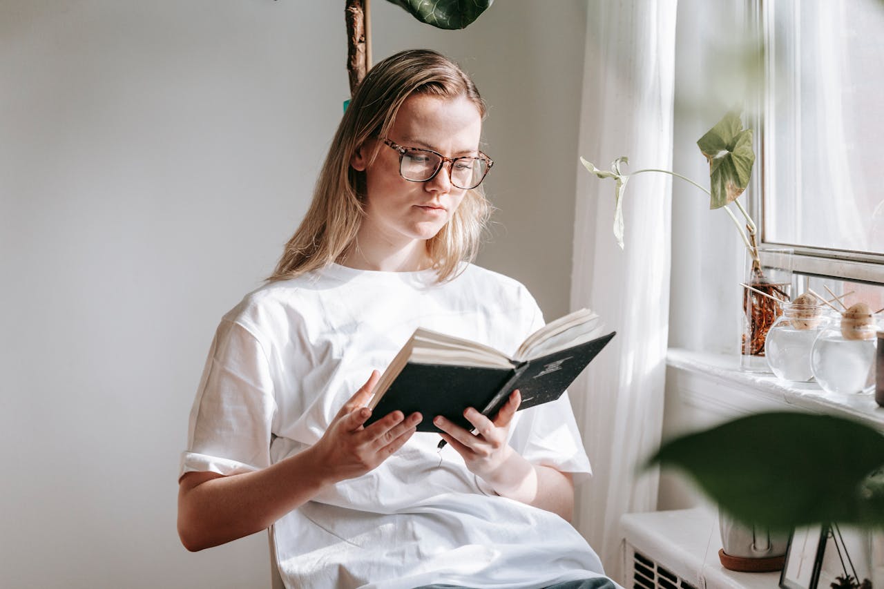 services-05 Young woman in glasses reading a book by a sunlit window. Calm and focused atmosphere.