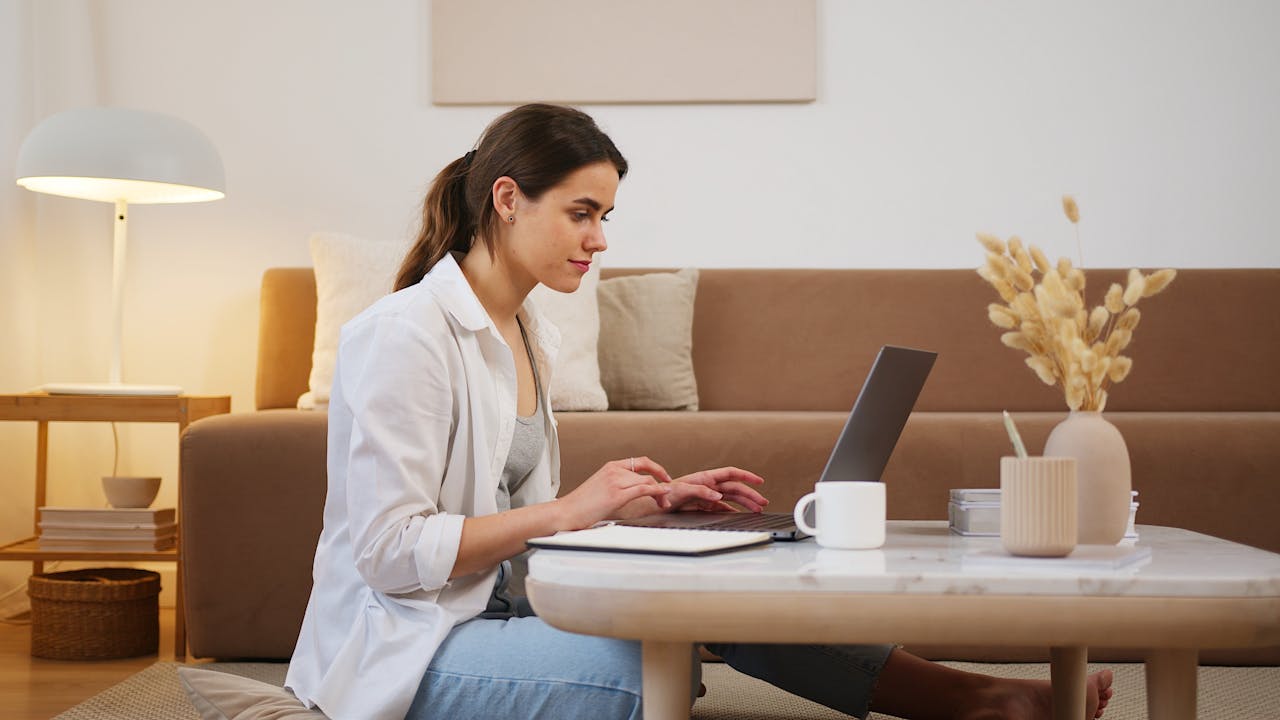 gallery-04 Side view of positive young lady with ponytail in casual outfit browsing contemporary netbook on coffee table while sitting on floor in cozy living room