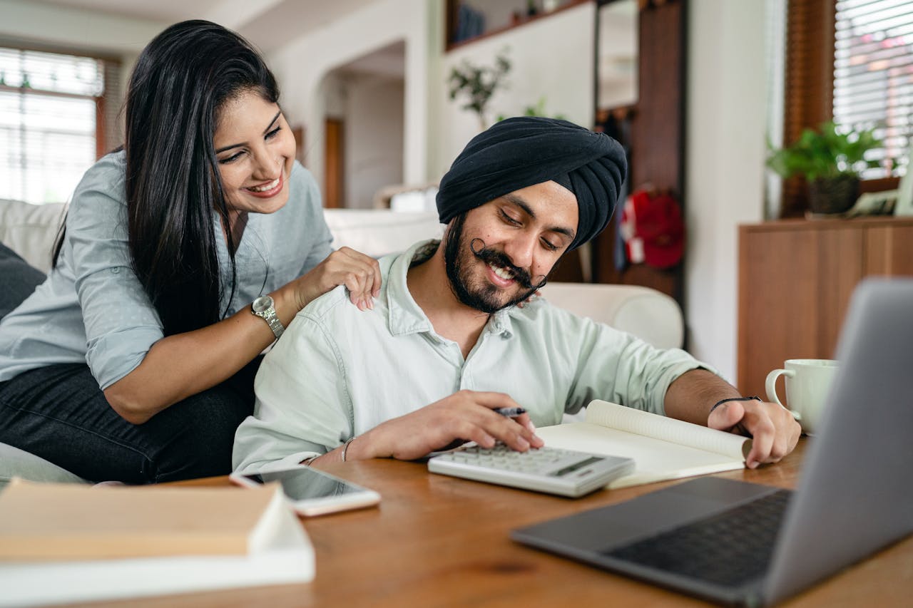 A smiling couple collaborates on financial paperwork at home, using a laptop and calculator.