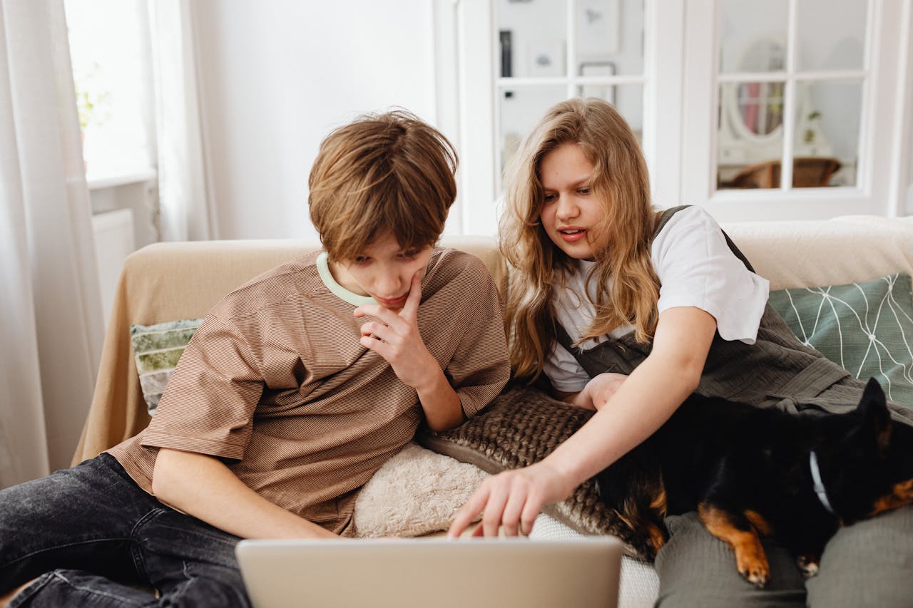 Two siblings with their dog enjoy quality time on a sofa using a laptop together.